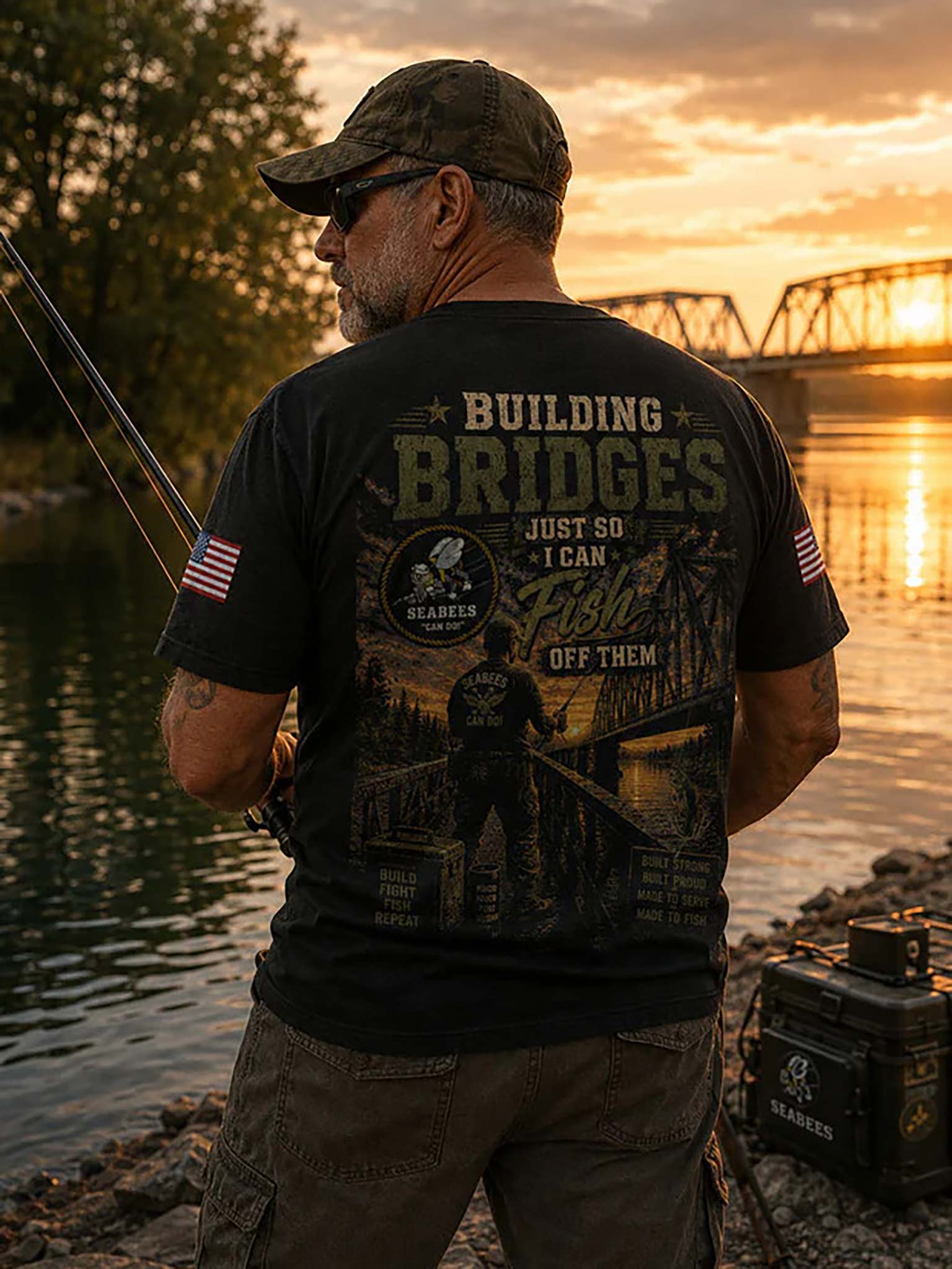 A man stands on a rocky riverbank at sunset, shown from the back, wearing a black graphic T-shirt with 'BUILDING BRIDGES JUST SO I CAN Fish OFF THEM' and a large fisherman illustration. A fishing rod is extended, and a branded black tackle box sits nearby on the shore.