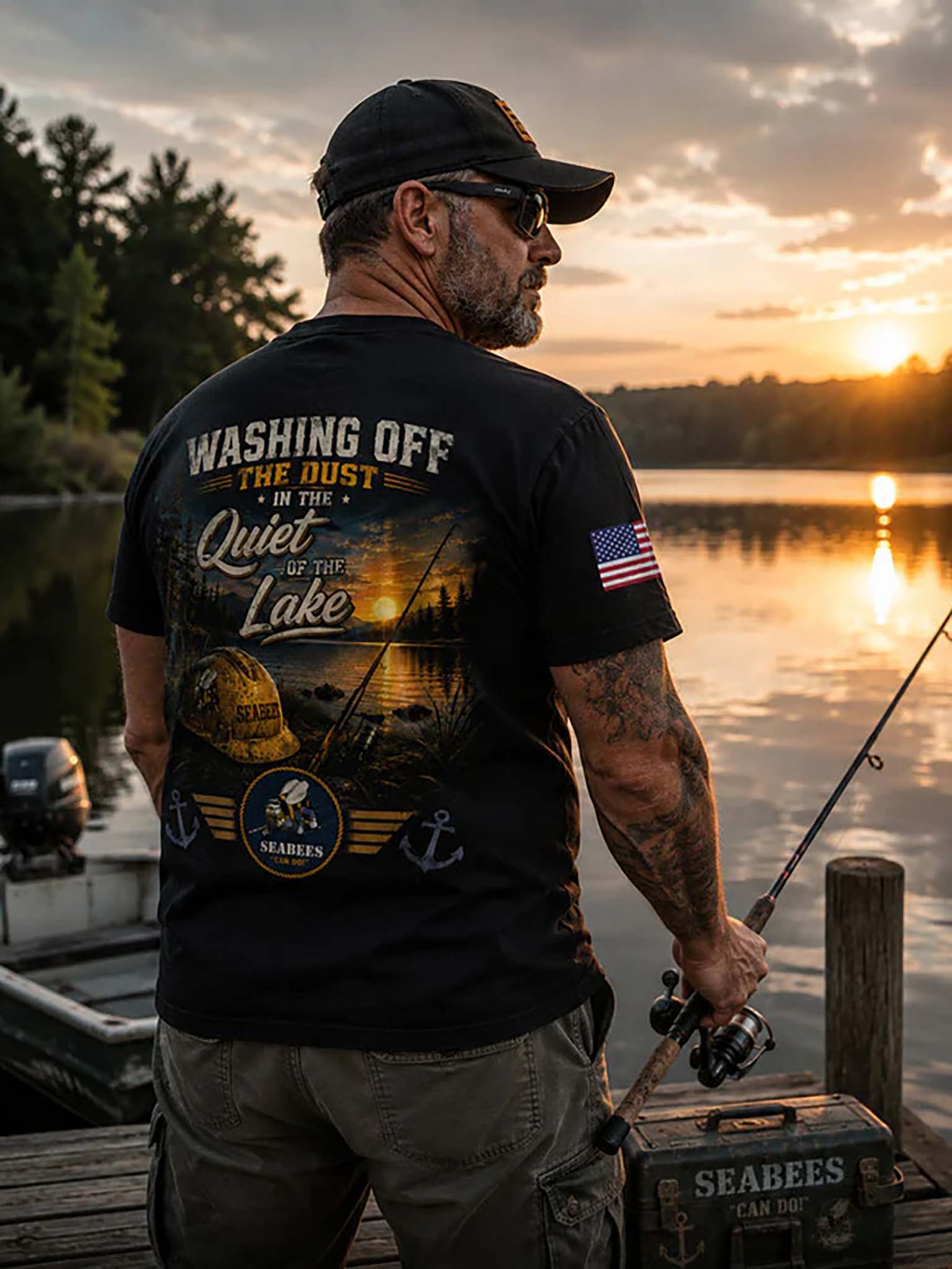 A man stands on a dock at sunset wearing a black graphic t-shirt with a large fishing-themed back print that reads 'WASHING OFF THE DUST IN THE Quiet OF THE Lake' and an American flag patch on the sleeve. He is holding a fishing rod next to a boat at dusk.