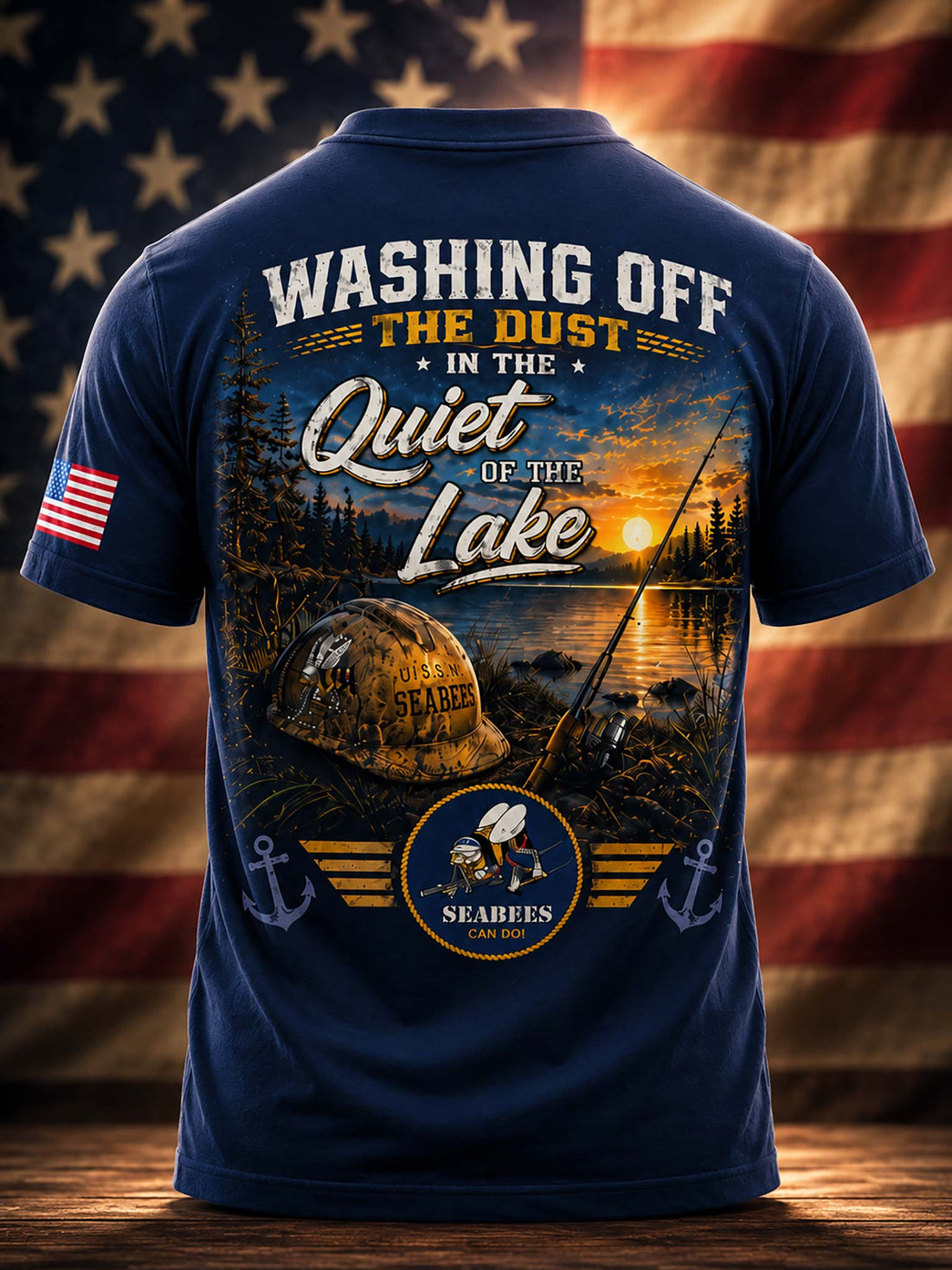 A black short-sleeve t-shirt shown from the back, featuring a large outdoor fishing graphic with the text 'WASHING OFF THE DUST IN THE Quiet OF THE Lake' and a sunset lake scene. The shirt is displayed on a wooden surface with a blurred American flag background.