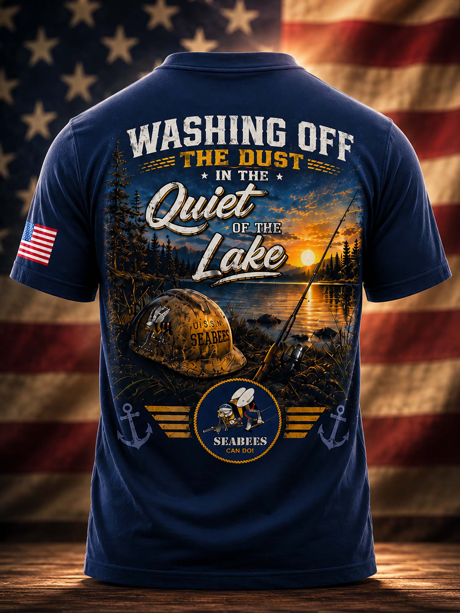 A navy blue short-sleeve t-shirt shown from the back, featuring a large graphic print with a lake sunset, a fishing helmet, a fishing rod, anchors, and the text 'WASHING OFF THE DUST IN THE Quiet OF THE Lake'. The shirt is displayed against a blurred American flag background.
