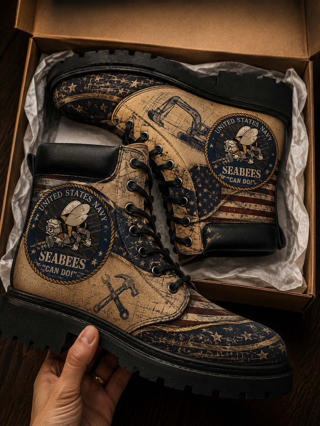 A pair of distressed military-style combat boots with a vintage US Navy Seabees print, displayed inside a brown cardboard box with tissue paper. A hand is holding the boot on the left side.