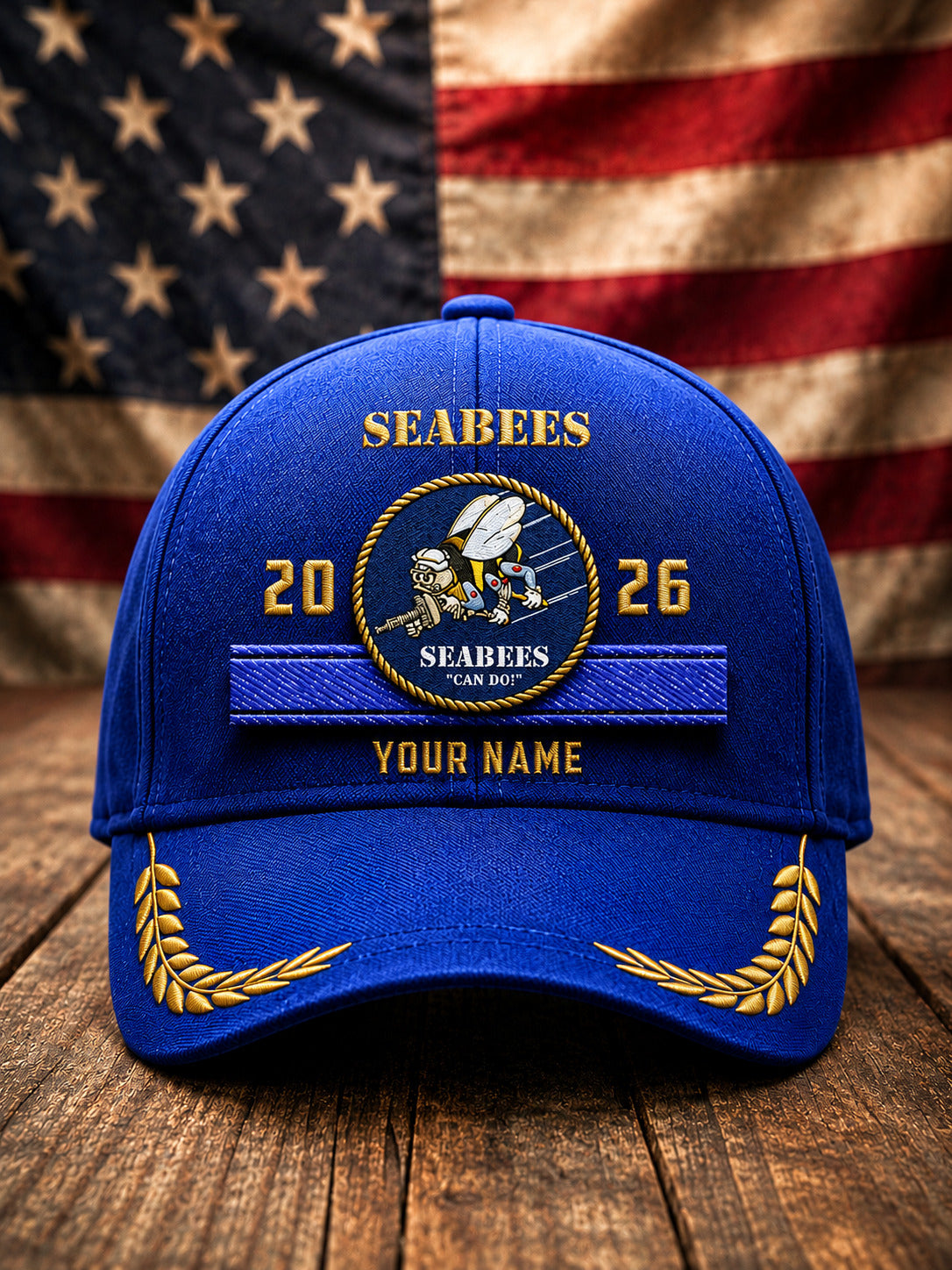 A royal blue baseball cap with gold and white embroidered Seabees insignia, featuring 'SEABEES', '2026', 'YOUR NAME', and laurel wreath embroidery on the brim, set against a rustic wooden surface with a blurred American flag background.