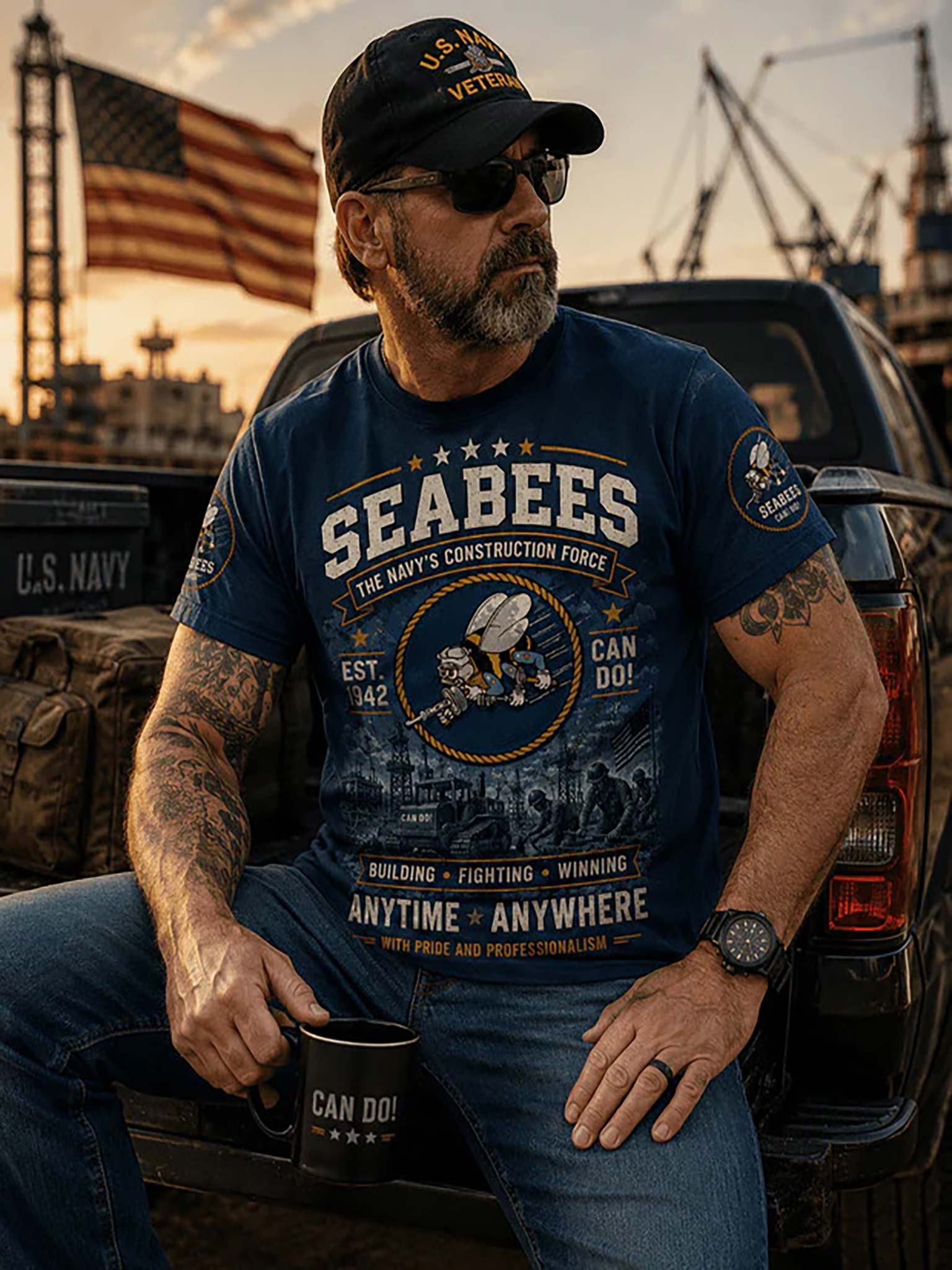A man wearing a navy blue graphic t-shirt with 'SEABEES' printed on the front, sitting in the bed of a truck at sunset. He is wearing a matching 'U.