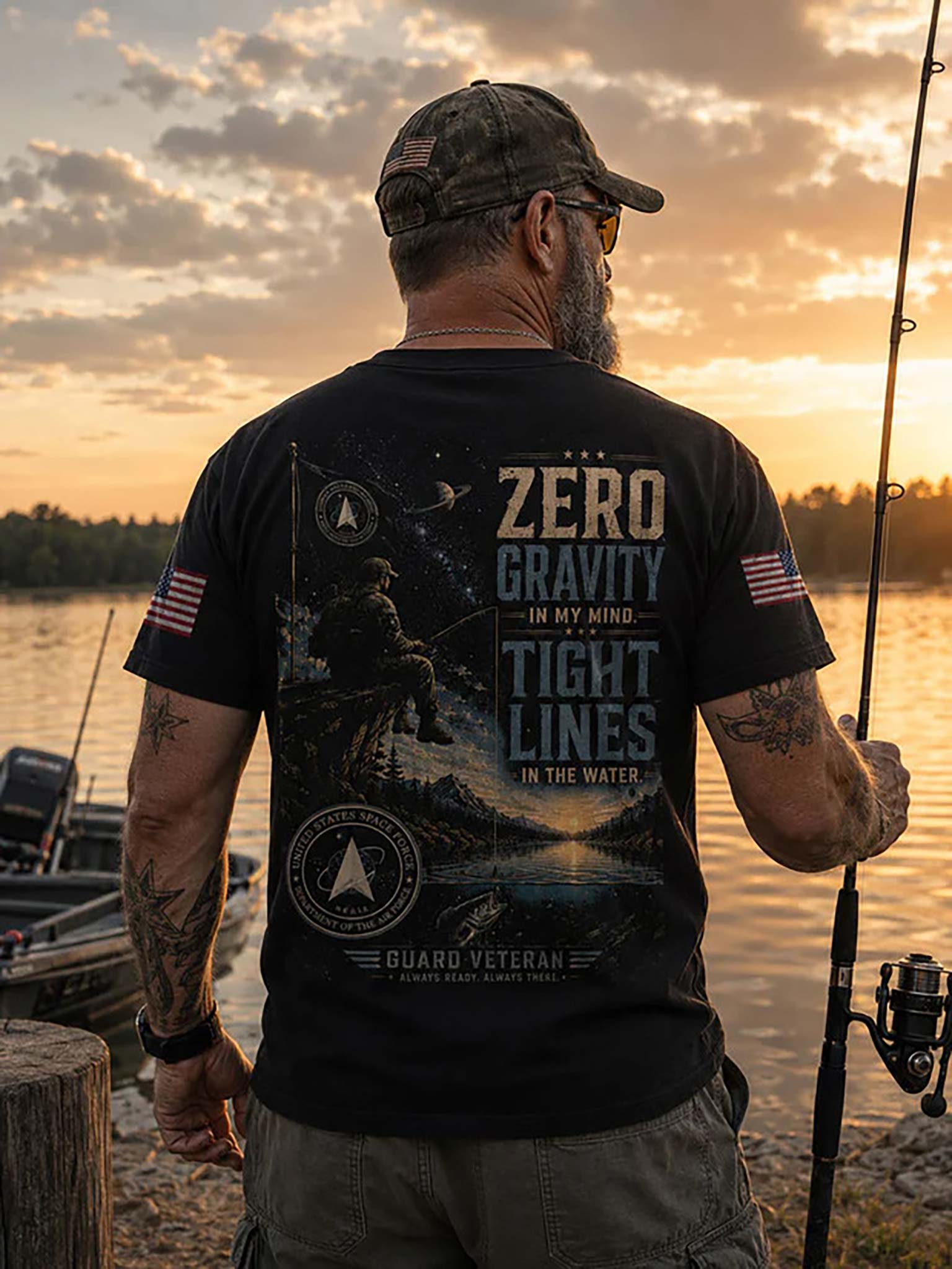 A man stands on a lakeshore at sunset wearing a black graphic T-shirt with patriotic and fishing-themed designs, including the text 'ZERO GRAVITY IN MY MIND TIGHT LINES IN THE WATER' and 'GUARO VETERAN.'.