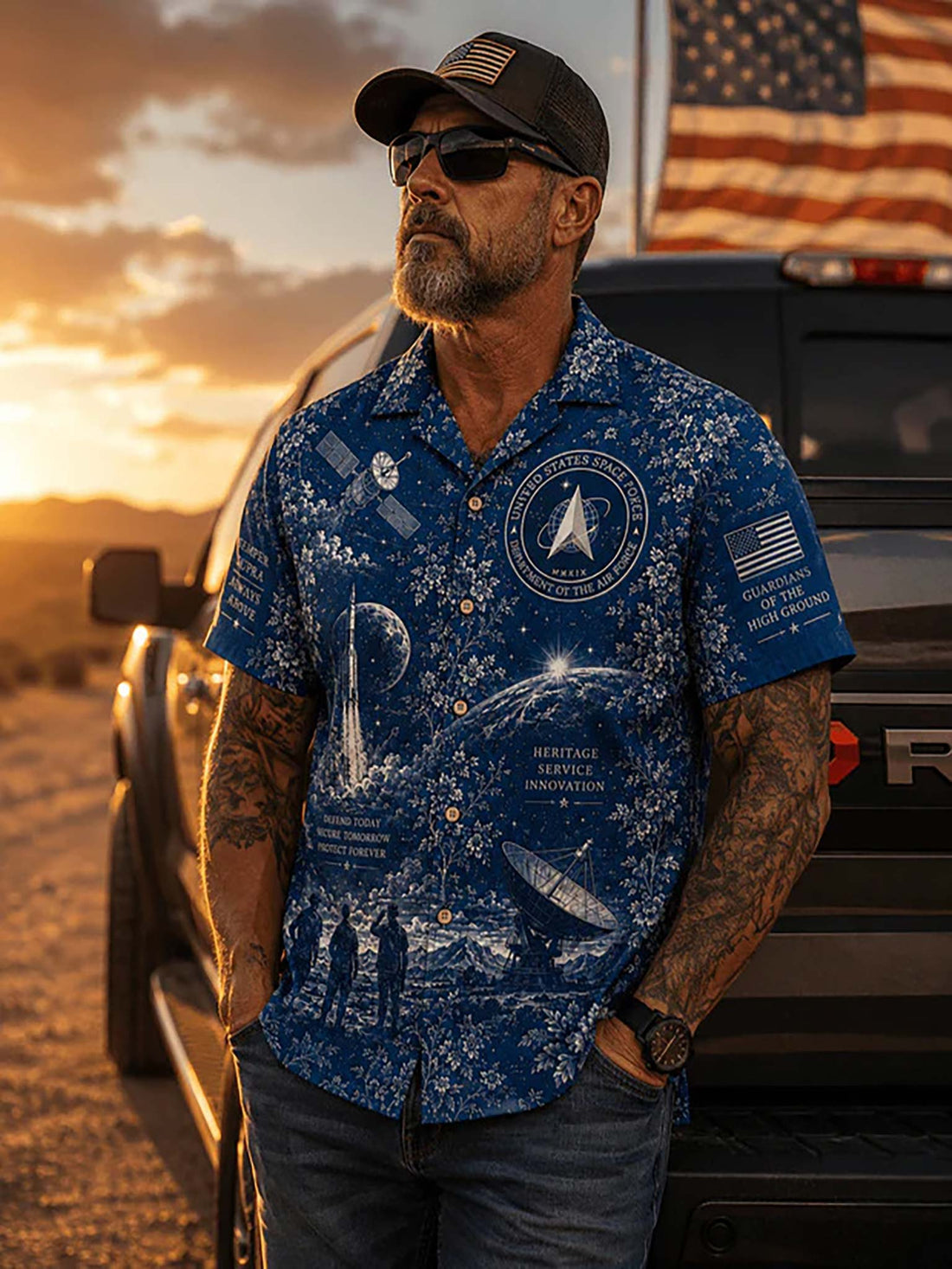 A blue short-sleeve button-up camp shirt with a white space-themed floral print, featuring illustrations of rockets, satellites, a space station, and military insignia. The shirt is displayed flat on a dark wood background with a matching blue cap, a folded American flag, and a glass of amber drink nearby.