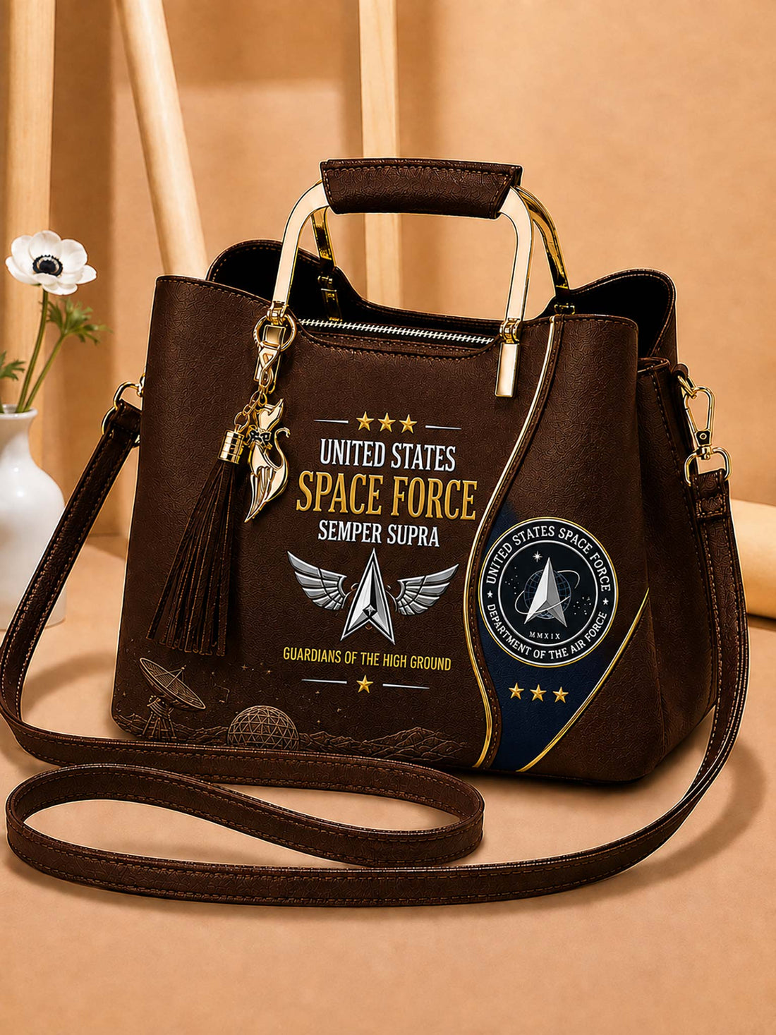 A dark brown faux-leather handbag with a top handle and shoulder strap, featuring a United States Space Force graphic with gold, white, and blue details, a tassel charm, and a gold-tone top handle.