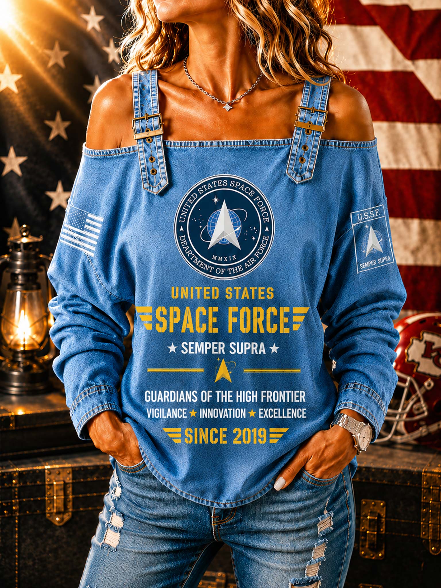 A woman wearing a blue off-the-shoulder long-sleeve top with United States Space Force graphics, paired with distressed blue jeans, posed in front of an American flag and a vintage lantern.