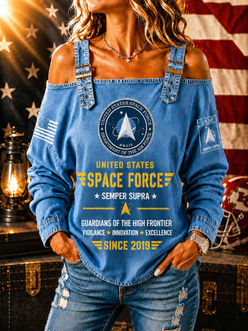 A woman wearing a blue off-the-shoulder long-sleeve top with United States Space Force graphics, paired with distressed blue jeans, posed in front of an American flag and a vintage lantern.