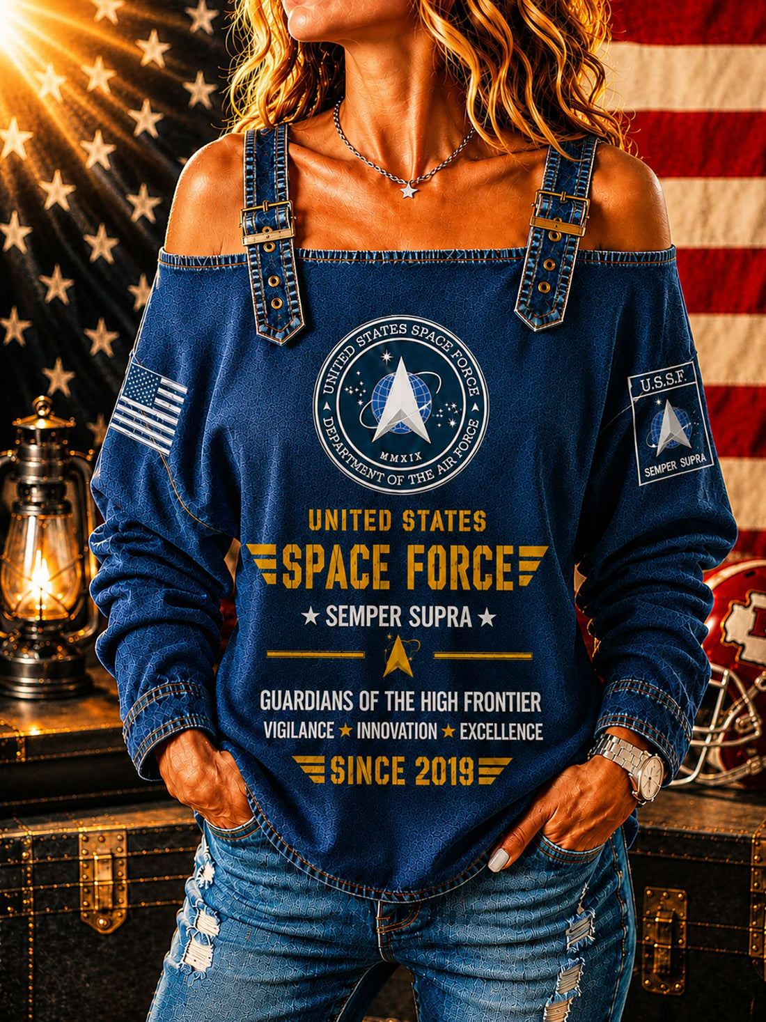 A woman wearing a blue off-the-shoulder long-sleeve top with United States Space Force graphics, paired with distressed blue jeans, posed in front of an American flag and a vintage lantern.