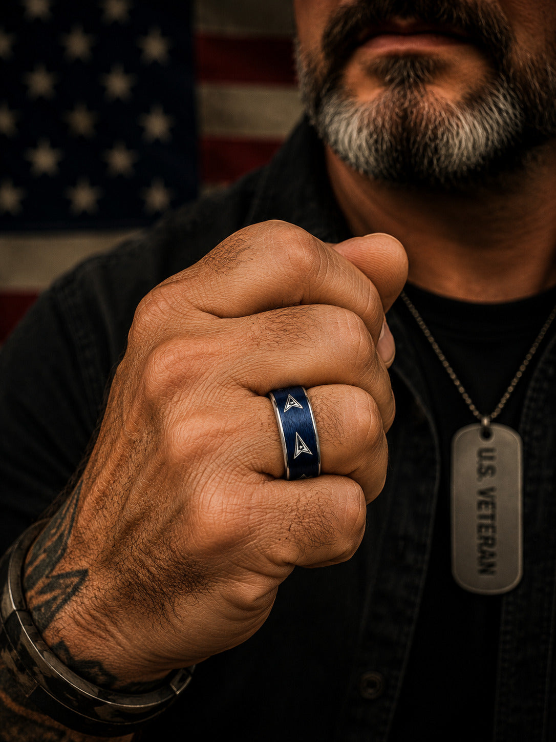 Two matching silver-toned wedding bands with brushed blue inlays and triangular insignia motifs, one engraved with 'SPACE FORCE,' photographed against a blurred American flag background.