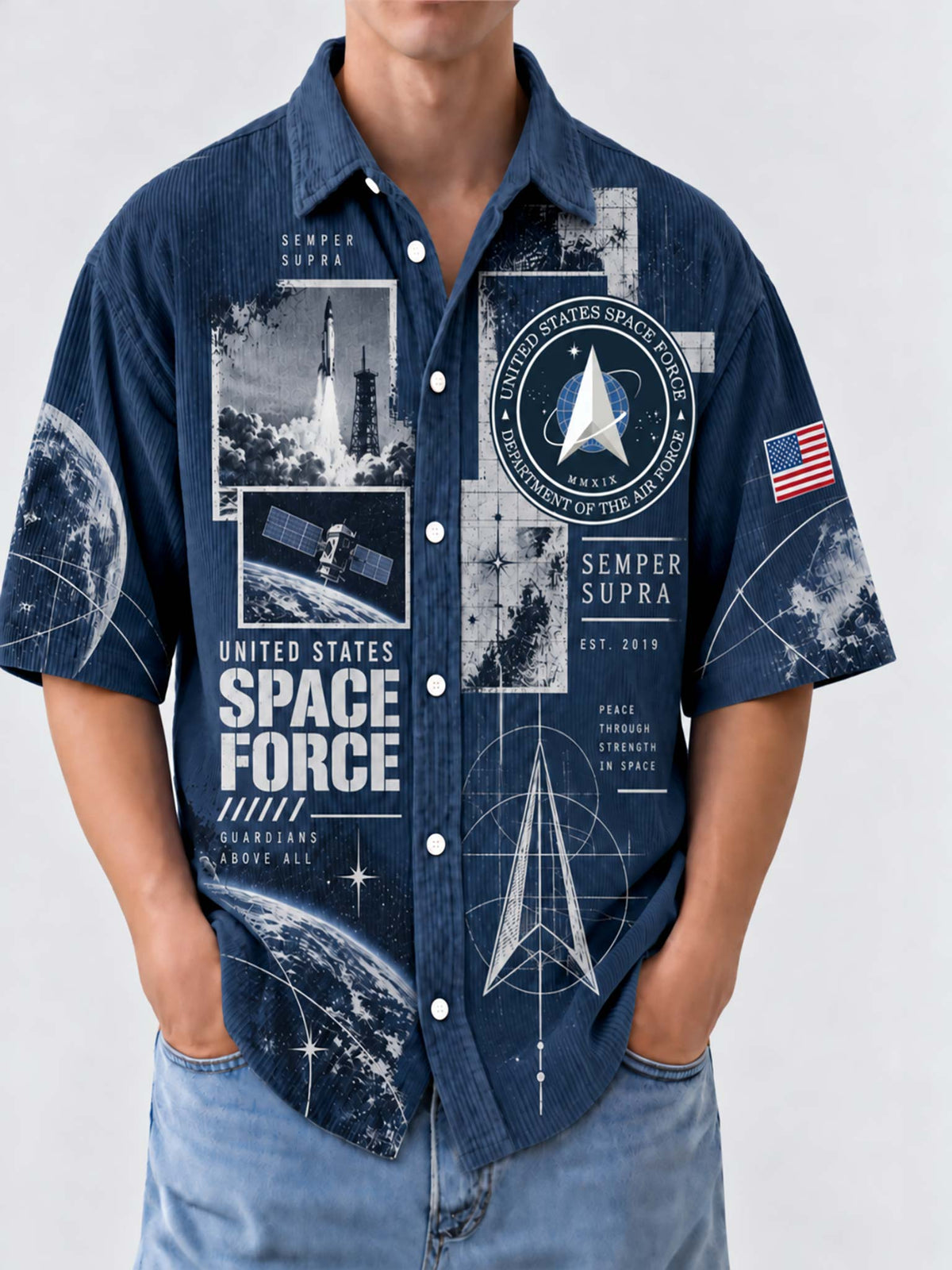 A man wearing a short-sleeve button-up shirt with a dark blue space-themed graphic print featuring 'United States Space Force' text, rockets, satellites, a delta symbol, and an American flag patch on the sleeve, paired with light blue jeans against a light gray background.