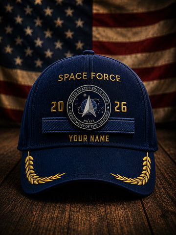 A navy blue baseball cap with gold embroidered details, featuring a 'Space Force' emblem on the front, displayed on a wooden surface with a blurred American flag in the background.