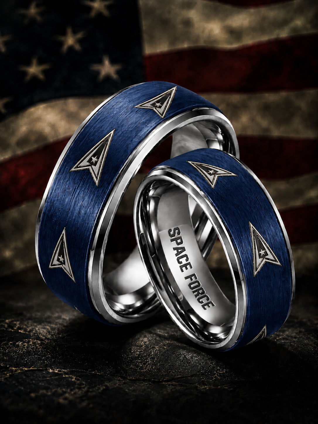 Two matching silver-toned wedding bands with brushed blue inlays and triangular insignia motifs, one engraved with 'SPACE FORCE,' photographed against a blurred American flag background.