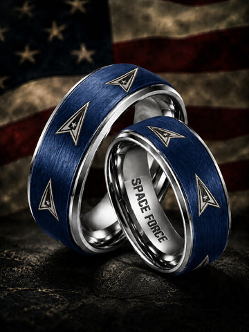 Two matching silver-toned wedding bands with brushed blue inlays and triangular insignia motifs, one engraved with 'SPACE FORCE,' photographed against a blurred American flag background.