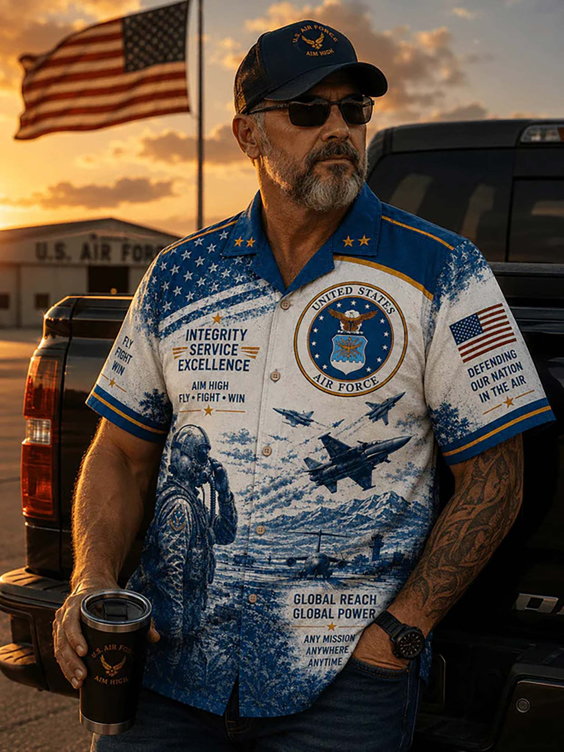 A short-sleeve button-up shirt with a blue and white United States Air Force theme, featuring military insignia, aircraft, a pilot silhouette, and patriotic text. The shirt is displayed on a dark wood surface with a matching cap, a US flag, a drink, and a plant leaf nearby.