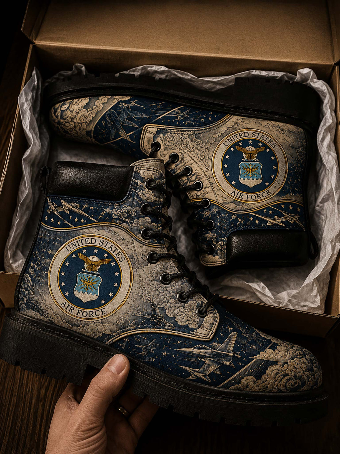 A pair of lace-up boots with a detailed United States Air Force theme, featuring the Air Force seal and fighter jets on a map background. The boots are shown inside a cardboard box with tissue paper, with a hand holding the front boot.