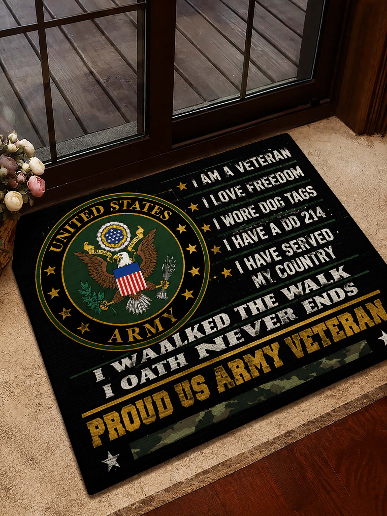 A rectangular black doormat with a large United States Army emblem and patriotic text, placed on a beige carpet in front of a glass door.