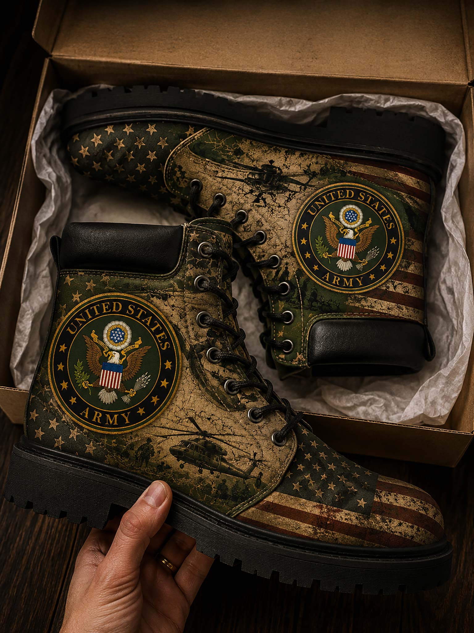 A pair of military-style boots with a distressed American flag and camouflage pattern, featuring a United States Army emblem on the side, displayed inside a cardboard shoebox with a hand holding one boot.