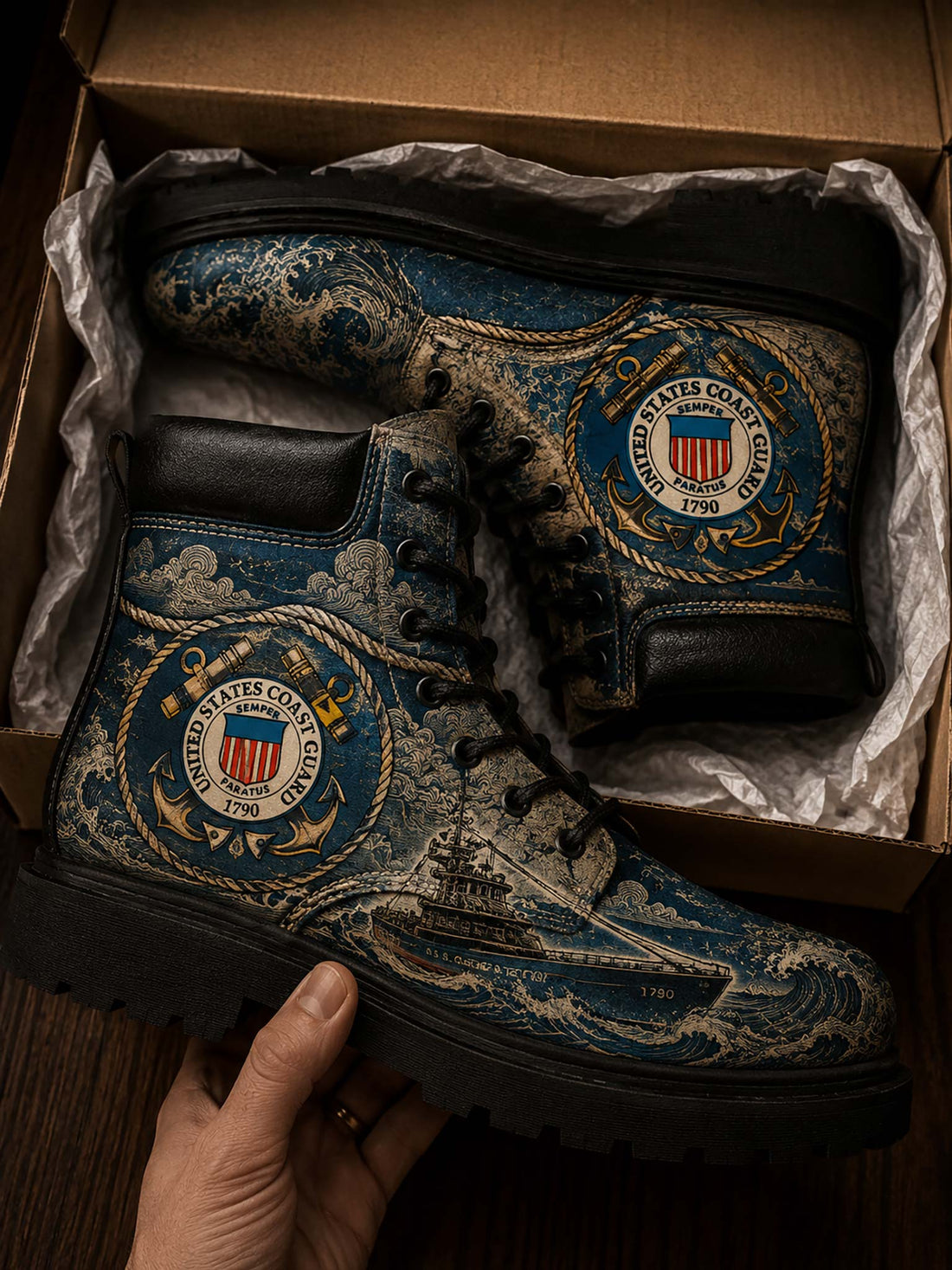 A pair of custom military-style boots with a blue and gold US Coast Guard wave pattern, displayed inside a cardboard box with a hand holding one boot.