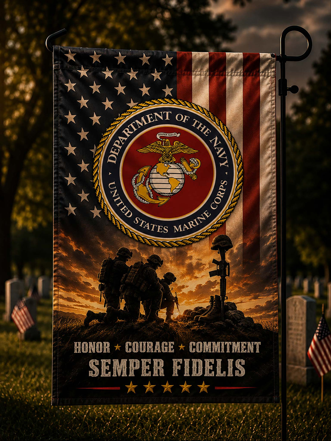 A vertical garden flag displayed on a black metal stand in a cemetery setting. The flag features the United States Marine Corps emblem over a stylized American flag and a sunset scene with soldiers and a headstone, with the text 'HONOR + COURAGE + COMMITMENT SEMPER FIDELIS' at the bottom.