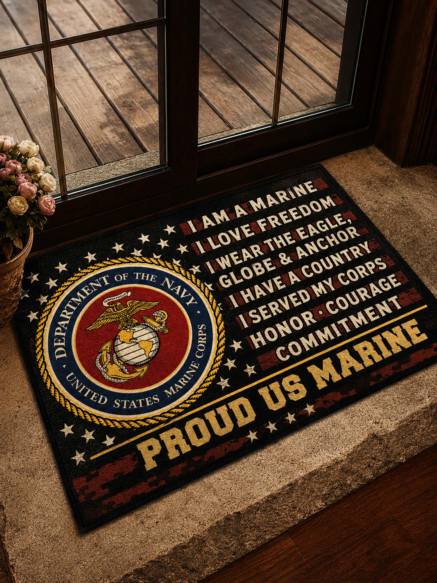 A rectangular doormat placed at a doorway, featuring the United States Marine Corps emblem on the left and printed text reading 'I AM A MARINE', 'I LOVE FREEDOM', 'I WEAR THE EAGLE, GLOBE & ANCHOR', 'I HAVE A COUNTRY', 'I SERVED MY CORPS', 'HONOR, COURAGE', and 'COMMITMENT', with 'PROUD US MARINE' across the bottom. The mat is shown in a real entryway setting with wood flooring, glass doors, and a small flower arrangement.
