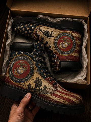 A pair of lace-up boots with a distressed American flag and United States Marine Corps theme, displayed inside a brown cardboard box with tissue paper. A hand is holding the boot at the bottom left.