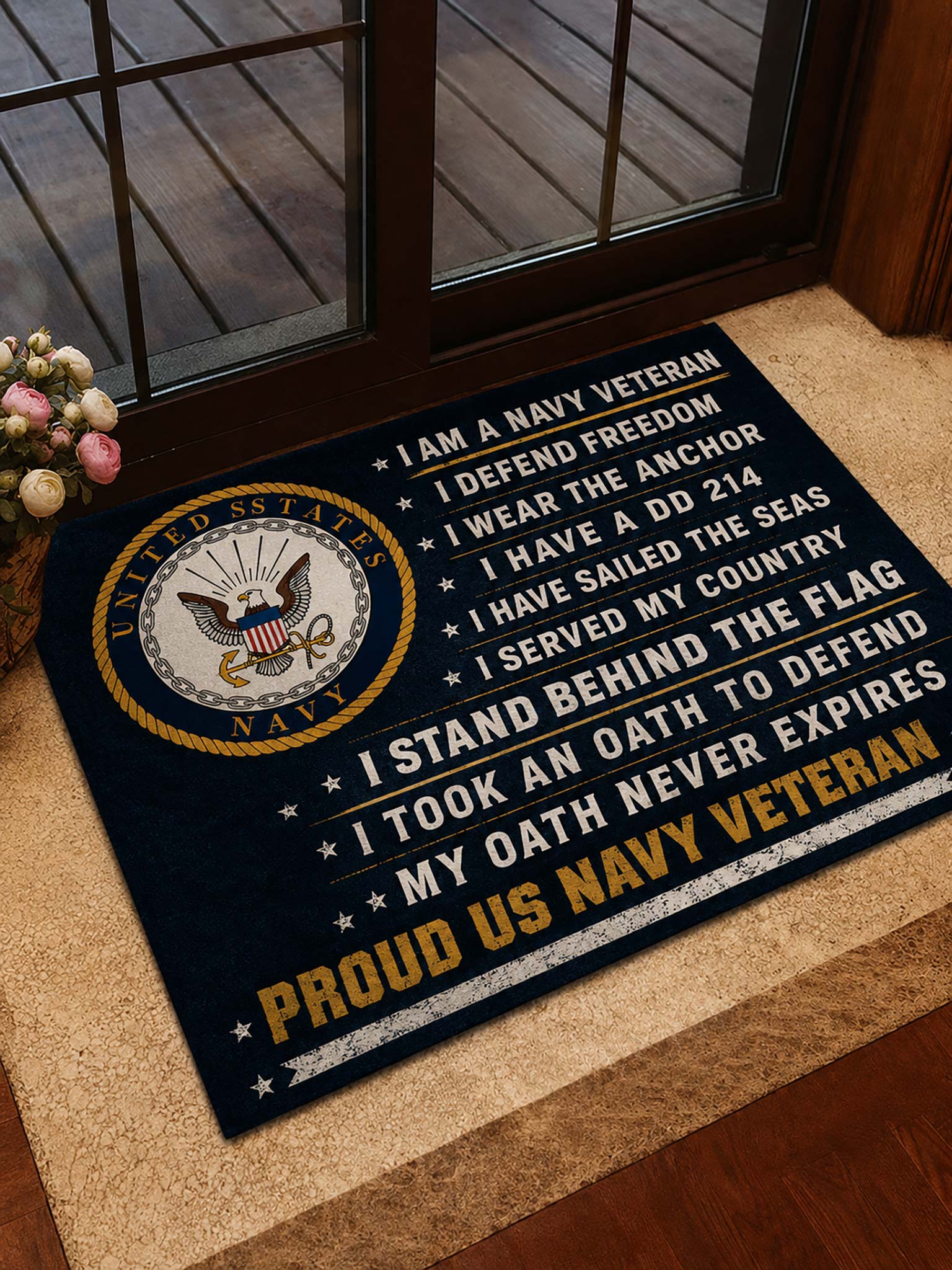 A navy blue doormat with a United States Navy emblem and patriotic text, placed on a tan entryway step in front of a glass door.