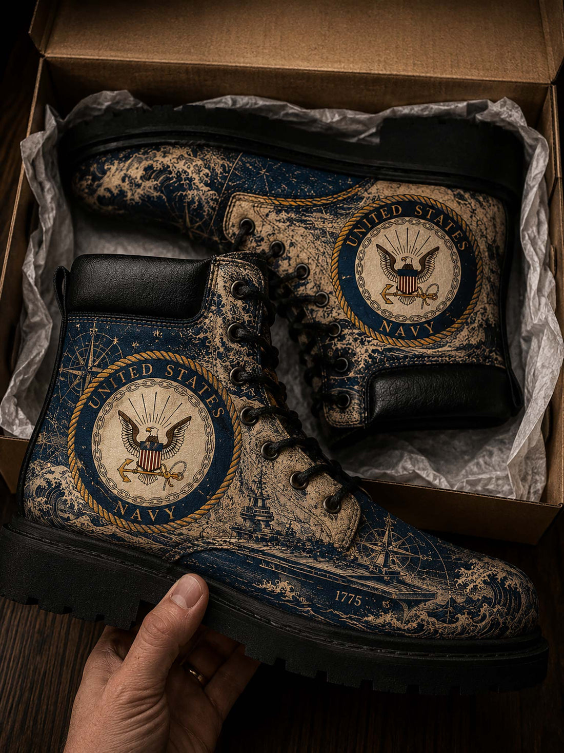 A pair of distressed navy blue and beige combat boots with United States Navy emblems, displayed inside a cardboard box with a hand holding one boot.