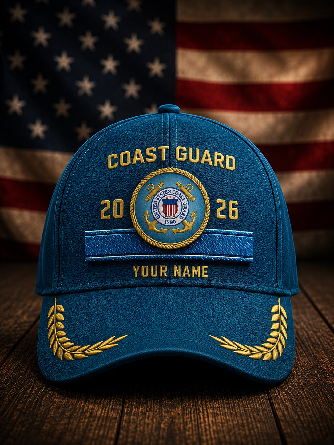 A blue Coast Guard themed baseball cap with gold embroidered details, including 'COAST GUARD', a central emblem, and 'YOUR NAME', displayed on a wooden surface with a blurred American flag in the background.