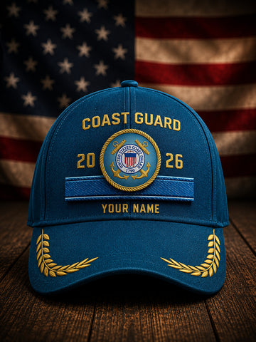 A blue Coast Guard themed baseball cap with gold embroidered details, including 'COAST GUARD', a central emblem, and 'YOUR NAME', displayed on a wooden surface with a blurred American flag in the background.