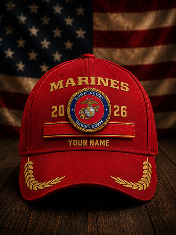 A red baseball cap with gold embroidered details, featuring a United States Marine Corps emblem, 'MARINES' text, and 'YOUR NAME' customization option, set against a blurred American flag and wooden background.