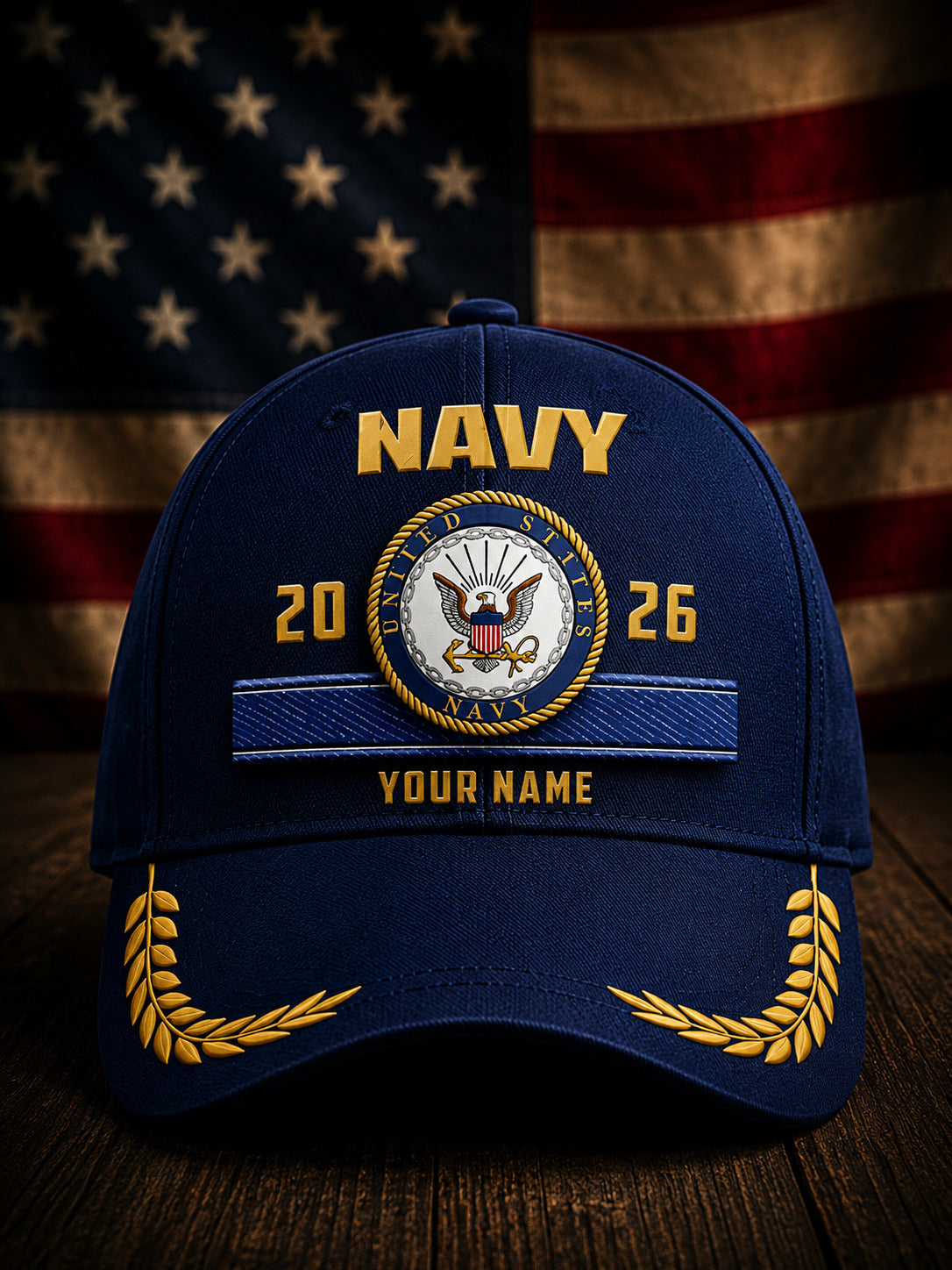 A navy blue baseball cap with gold and white embroidered details, featuring the word 'NAVY' and a custom emblem with the years '2026' and placeholder text 'YOUR NAME'. The cap is displayed on a wooden surface with a blurred American flag in the background.