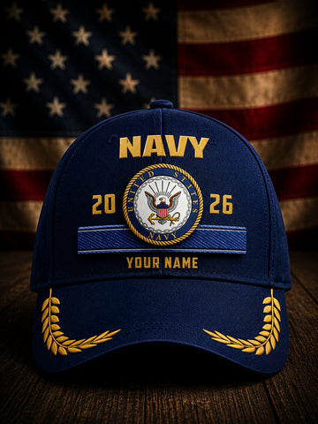 A navy blue baseball cap with gold and white embroidered details, featuring the word 'NAVY' and a custom emblem with the years '2026' and placeholder text 'YOUR NAME'. The cap is displayed on a wooden surface with a blurred American flag in the background.