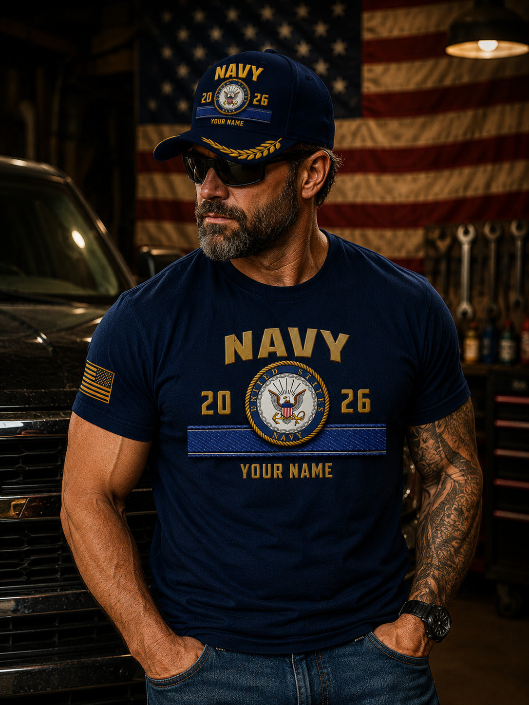 A navy blue baseball cap with gold and white embroidered details, featuring the word 'NAVY' and a custom emblem with the years '2026' and placeholder text 'YOUR NAME'. The cap is displayed on a wooden surface with a blurred American flag in the background.