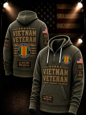 Two olive green quarter-zip hoodies shown from front and back, both printed with large 'VIETNAM VETERAN' text, a circular Vietnam Veterans emblem with red, yellow, green, and white stripes, an American flag patch, and a small 'EST 1955' patch. The back view is on the left and the front view is on the right, set against a dark patriotic background with spotlight effects and an American flag.