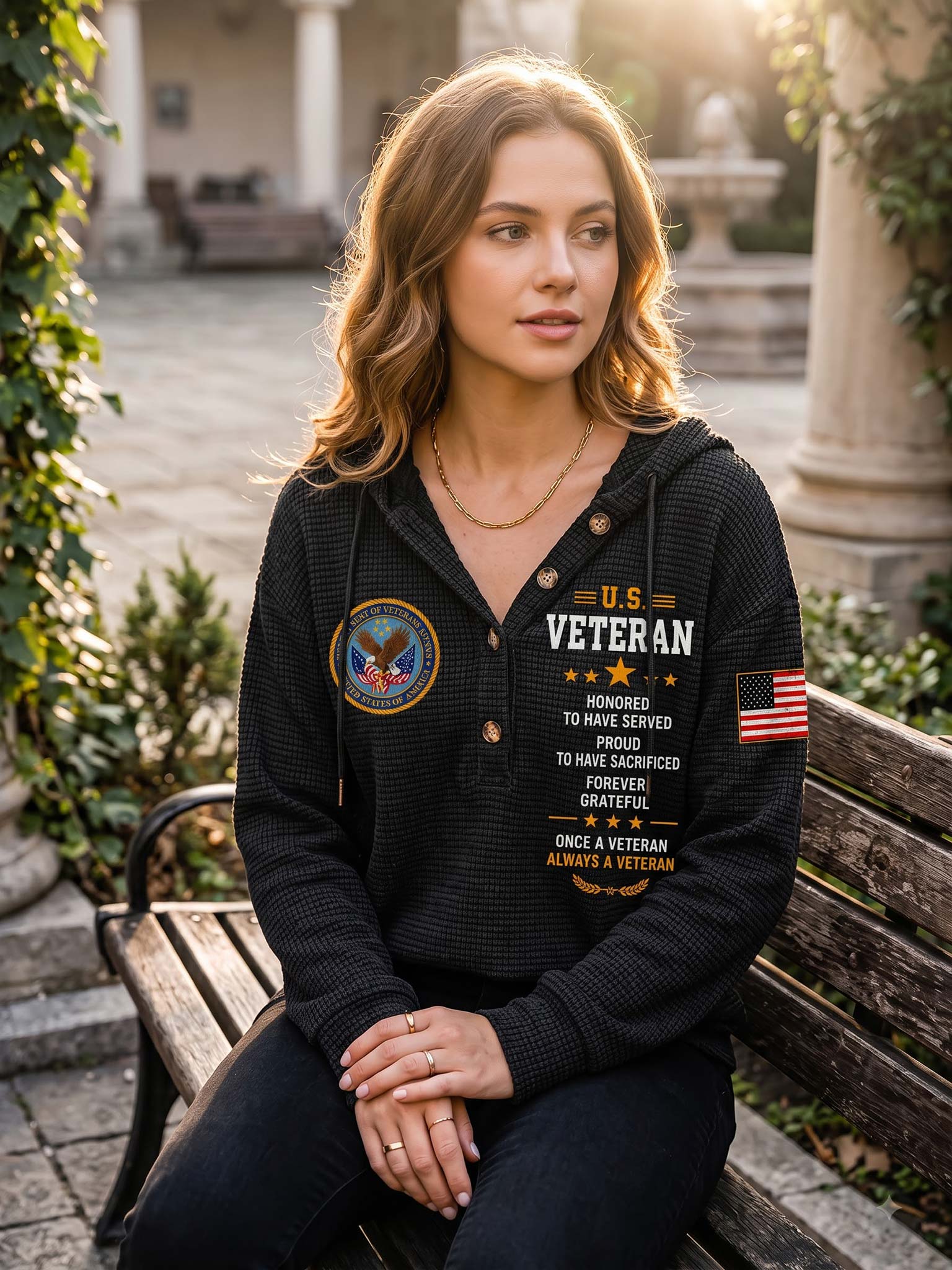 A woman sits on a wooden bench in an outdoor courtyard wearing a black waffle-knit hoodie with military-themed graphics, including a US Veteran emblem, a circular patch, and an American flag on the sleeve.