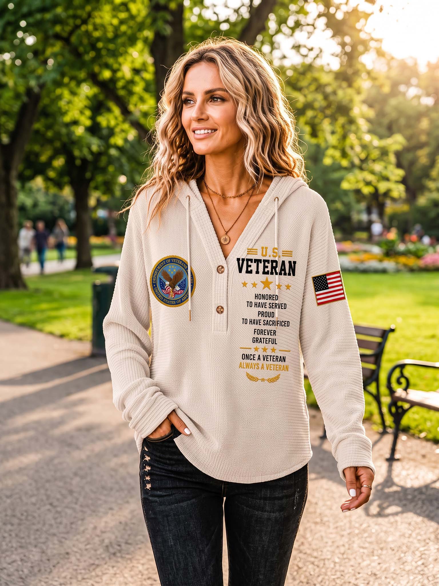 A woman wearing a beige waffle-knit hoodie with a 'U.S. VETERAN' graphic print on the front, standing in a sunny park.