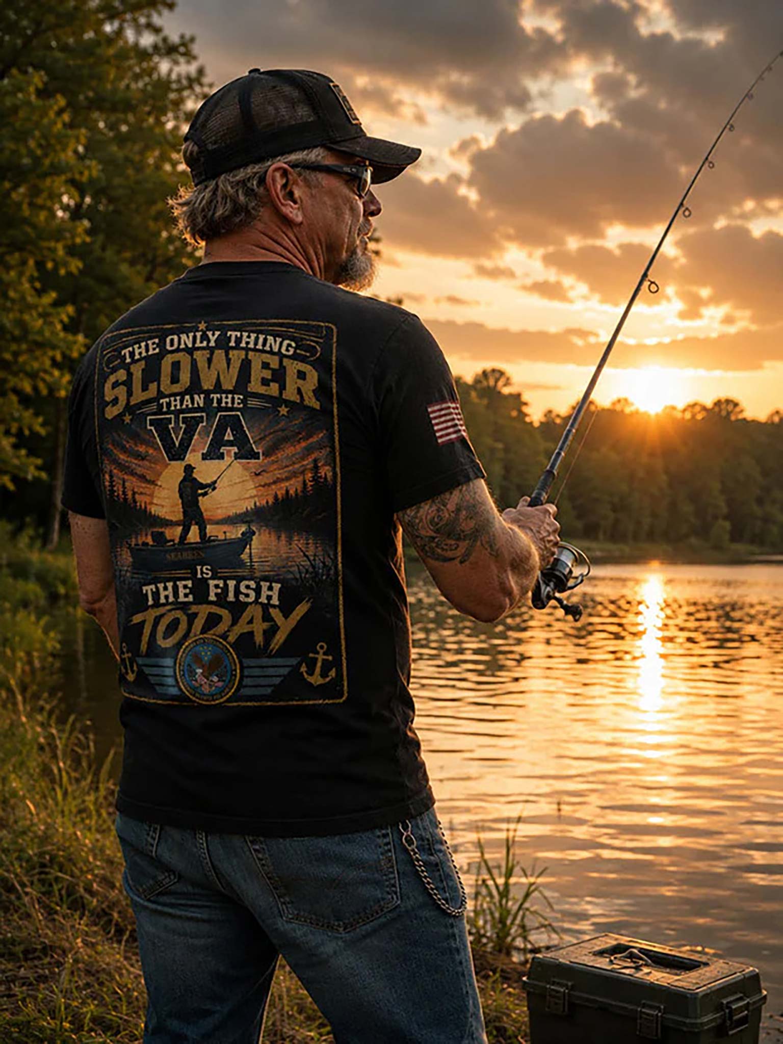 A man stands by a lake at sunset wearing a black graphic T-shirt with a large back print that reads 'THE ONLY THING SLOWER THAN THE VA IS THE FISH TODAY,' along with blue jeans and a fishing rod in hand.