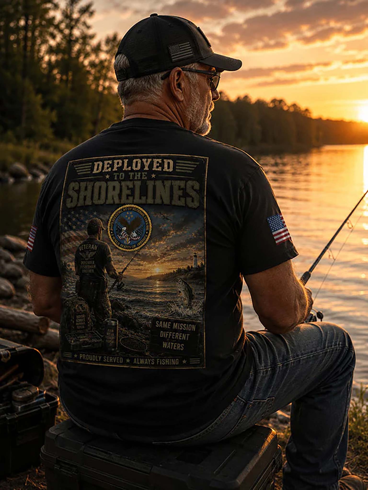 A man sitting by a lake at sunset wearing a black graphic T-shirt with a large back print reading 'Deployed to the Shorelines' and an American flag patch on the sleeve, seen from behind.