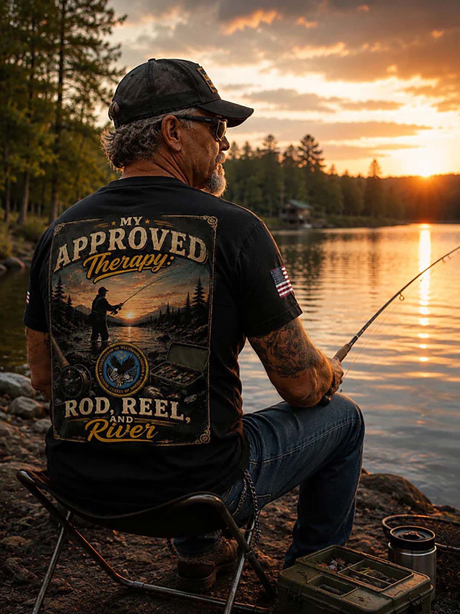 A man sits on a folding chair by a lake at sunset, holding a fishing rod. He is wearing a black graphic T-shirt with a large back print that reads 'My Approved Therapy: Rod, Reel, and River' over a scenic fishing illustration.