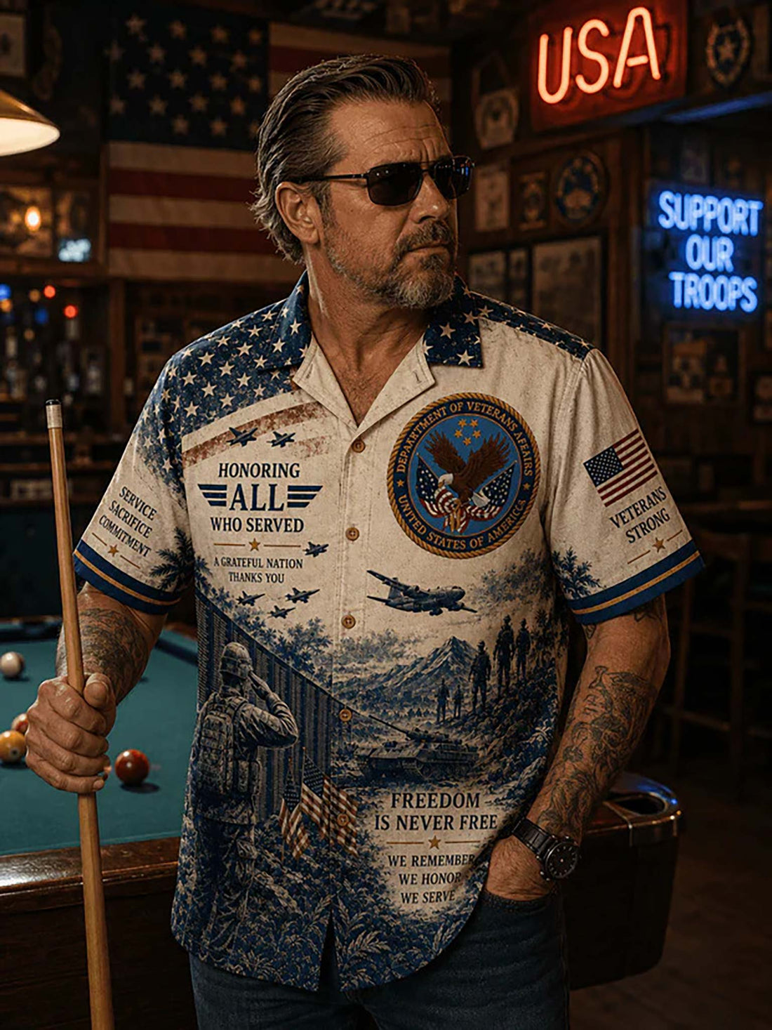 A short-sleeve button-up camp shirt with a patriotic military theme, featuring a blue and white American flag pattern, a soldier saluting, military aircraft and vehicles, and text including 'HONORING ALL WHO SERVED' and 'VETERANS STRONG'. The shirt is displayed on a dark wood surface with a blue cap, dog tags, a drink, and an American flag nearby.