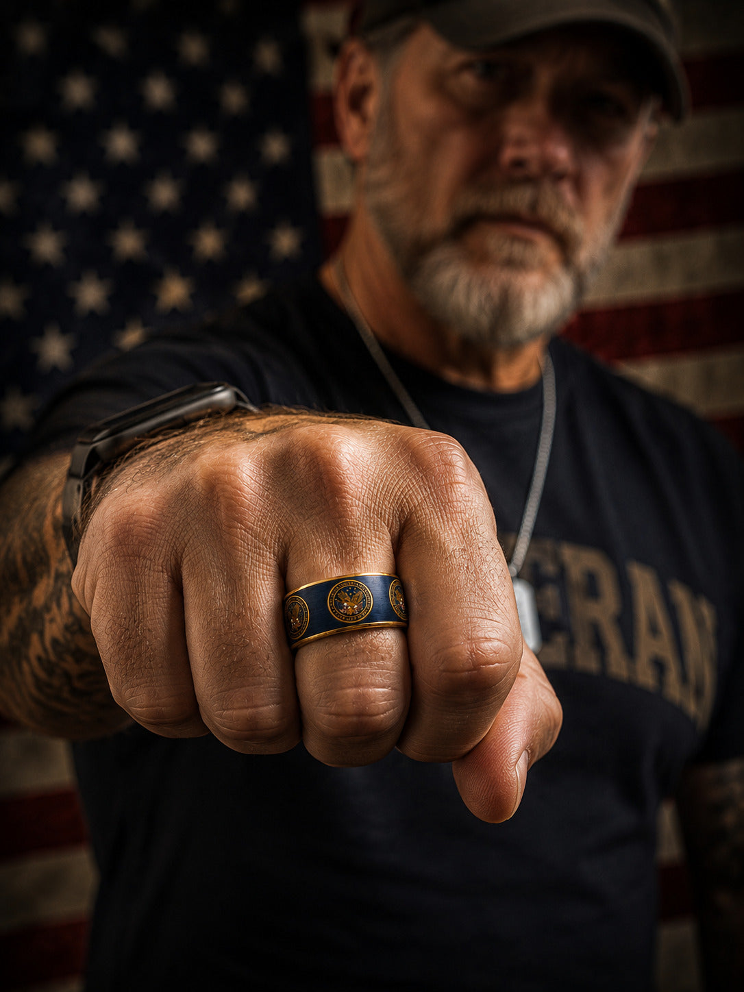 Two matching military-style rings with blue enamel, gold trim, and circular gold insignia, shown overlapping on a dark surface with an American flag blurred in the background. The inner band is engraved with the word "VETERAN.