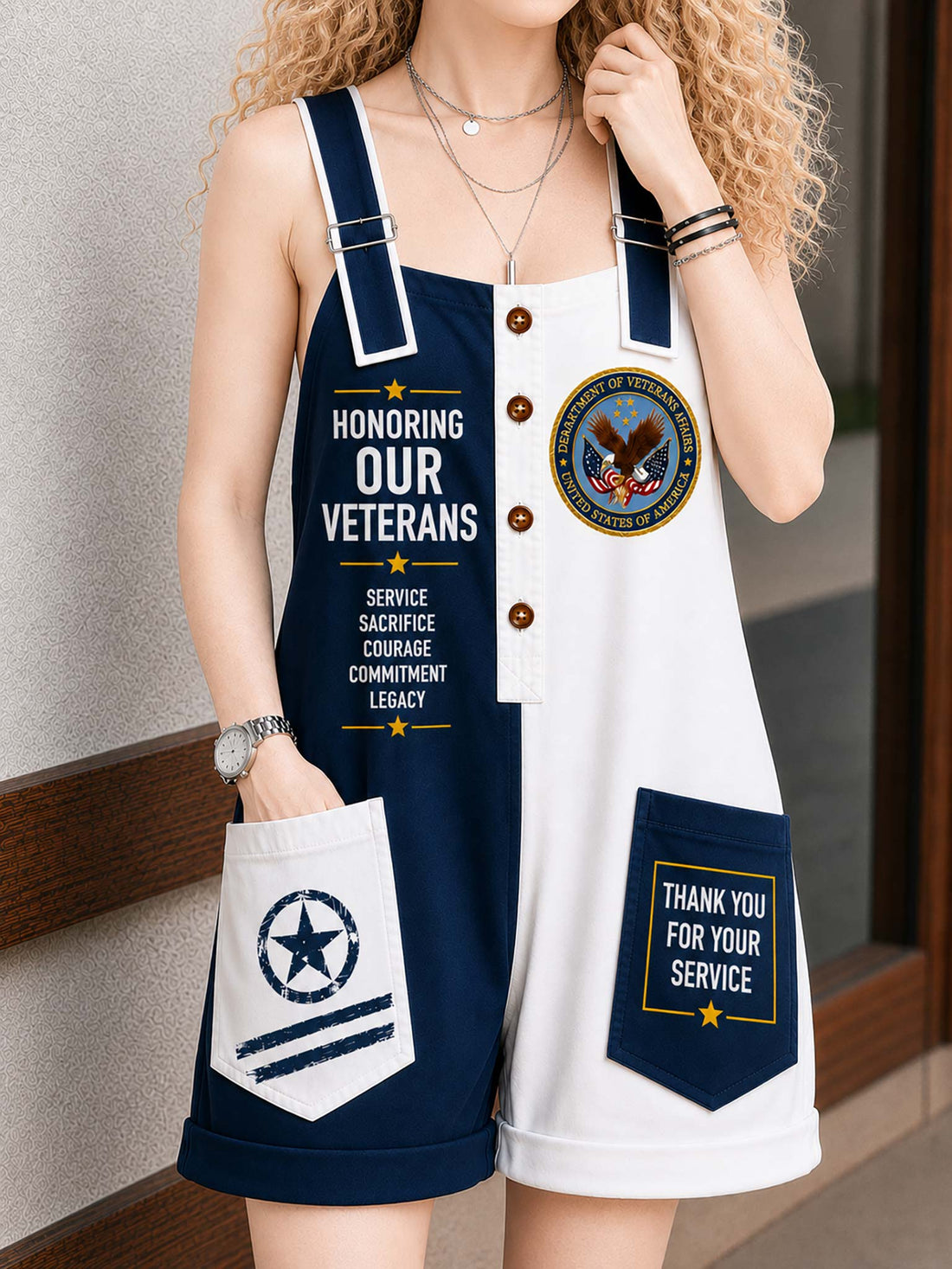 A woman wearing a navy and white short overall romper with patriotic military-themed graphics, including 'HONORING OUR VETERANS' text, a military seal, and a pocket that reads 'THANK YOU FOR YOUR SERVICE'.