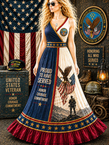 A woman wearing a patriotic military-themed maxi dress with a split design featuring navy blue and cream sections, stars, eagles, and text reading 'Proud to Have Served' and 'Honor Courage Commitment.' The scene is styled with an American flag, military medals, a lantern, and a wooden crate in a rustic indoor setting.