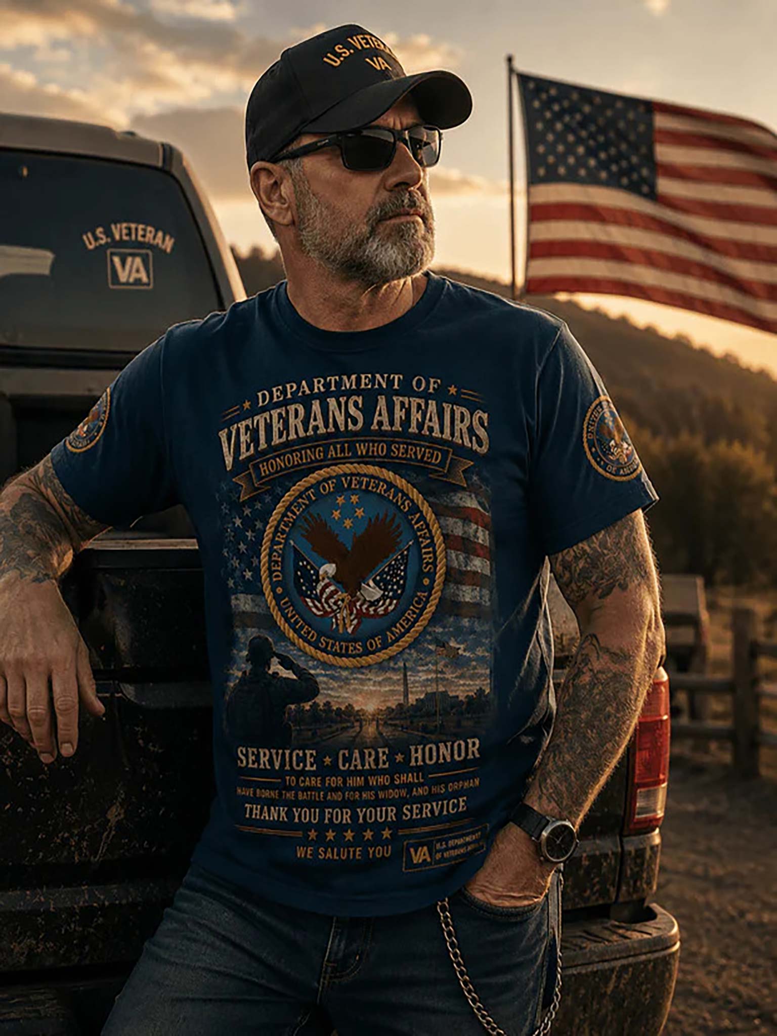 A man wearing a navy blue graphic t-shirt with 'Veterans Affairs' text and an eagle emblem, standing outdoors next to a truck with an American flag in the background.