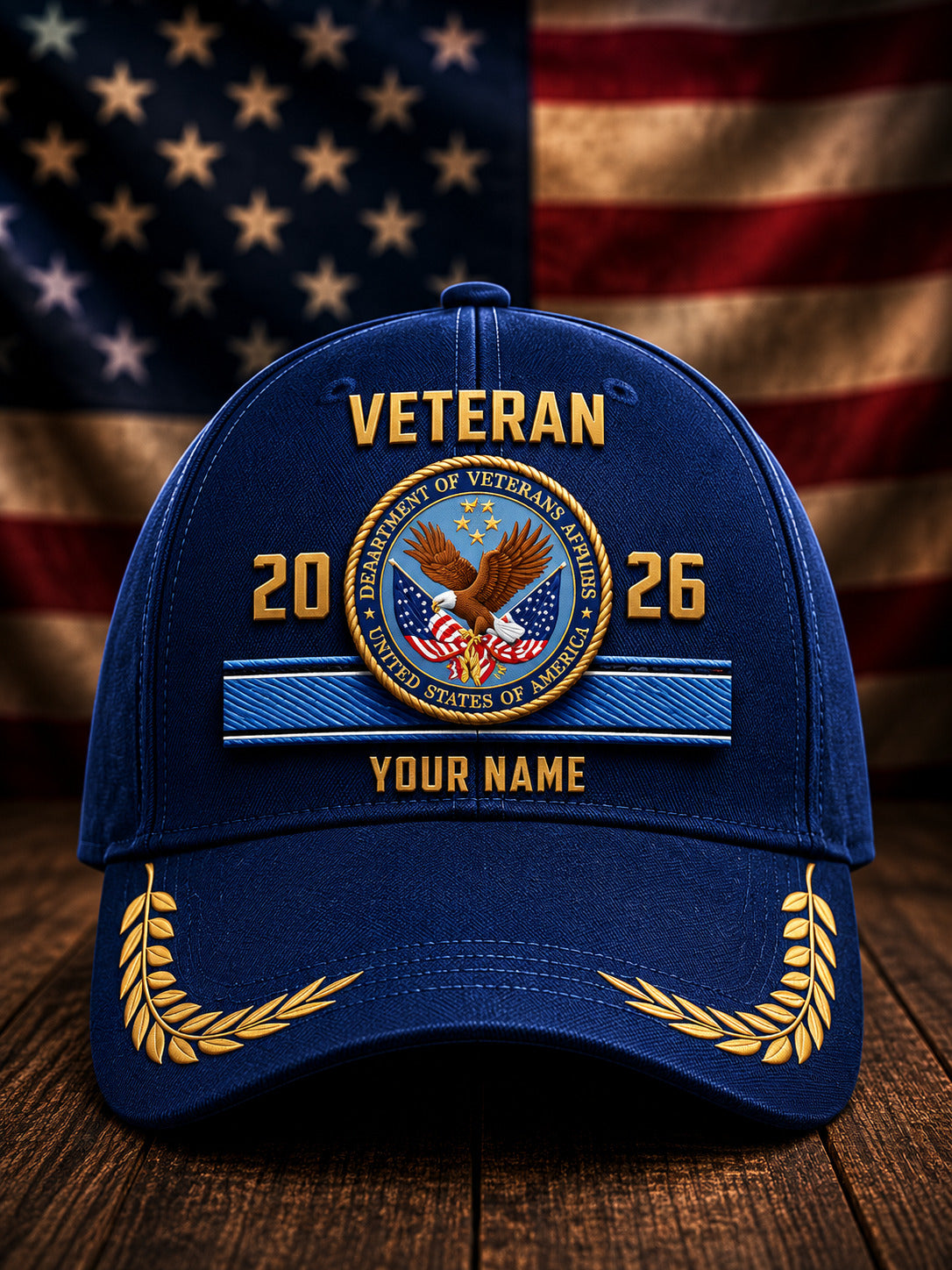 A royal blue baseball cap with a custom veteran design, featuring a gold and blue Department of Veterans Affairs seal, the text 'VETERAN 2026 YOUR NAME', and gold laurel wreaths on the brim, set against a blurred American flag and wooden table background.
