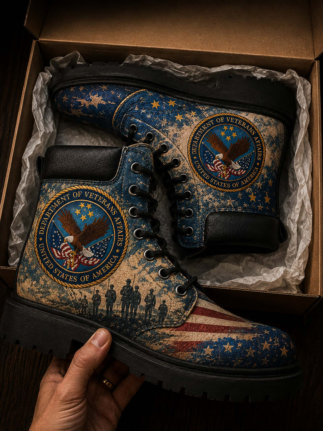 A pair of distressed military-style boots with a patriotic American flag and eagle design, displayed inside a brown shoebox with tissue paper, with a hand holding one boot.