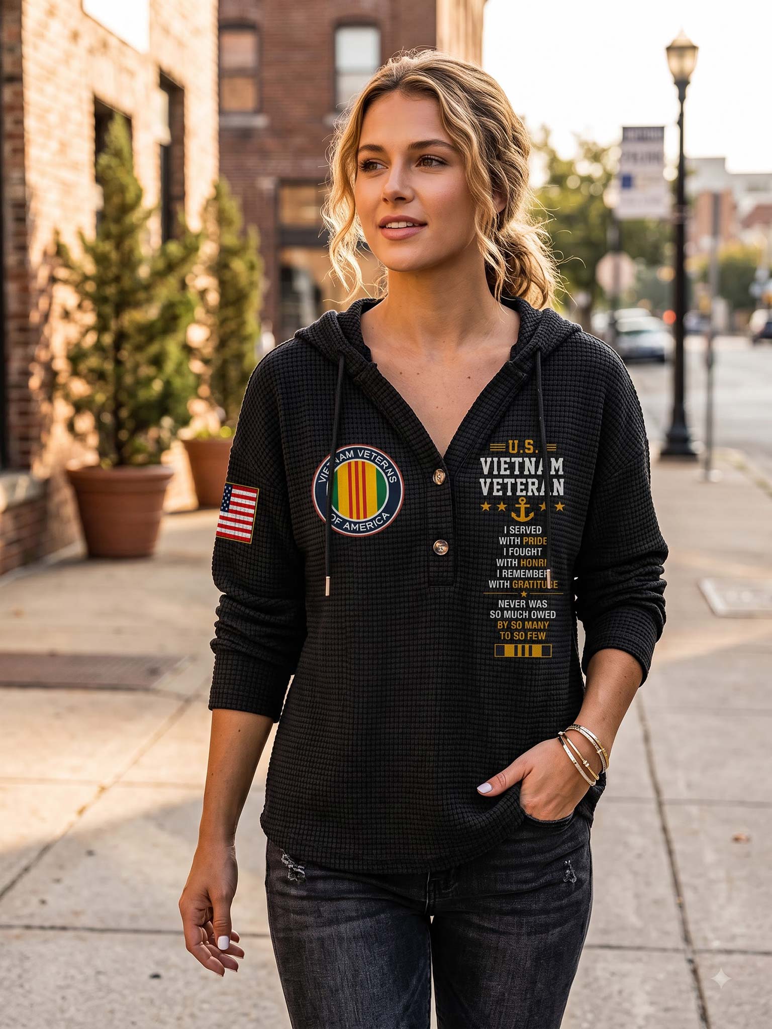 A woman stands on a city sidewalk wearing a black waffle-knit hoodie with military-themed graphics, including a Vietnam Veteran emblem and text, paired with dark jeans.