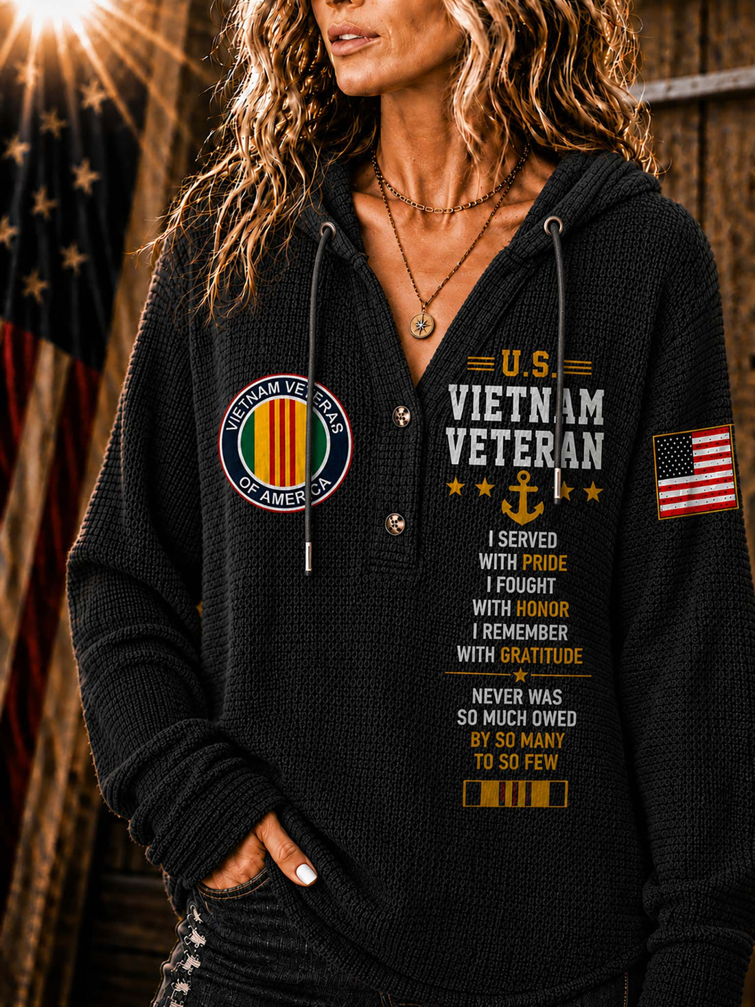 A woman wearing a beige ribbed hoodie with 'Vietnam Veteran' text, military-style patches, and an American flag graphic on the sleeve, posed against a rustic wooden background with a blurred American flag.