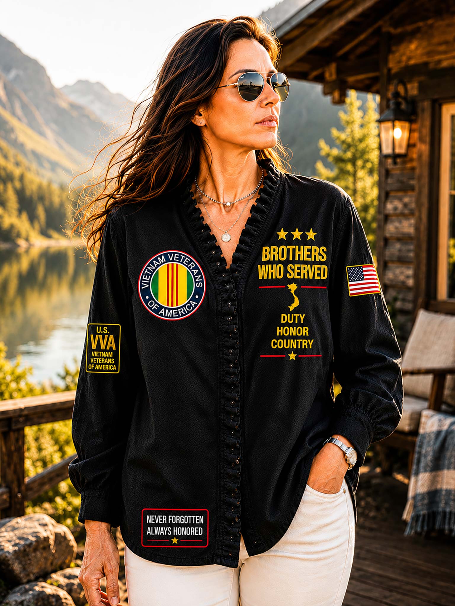 A woman wearing a black button-front shirt jacket with patriotic military-themed patches, standing on a wooden porch with mountains and a lake in the background.